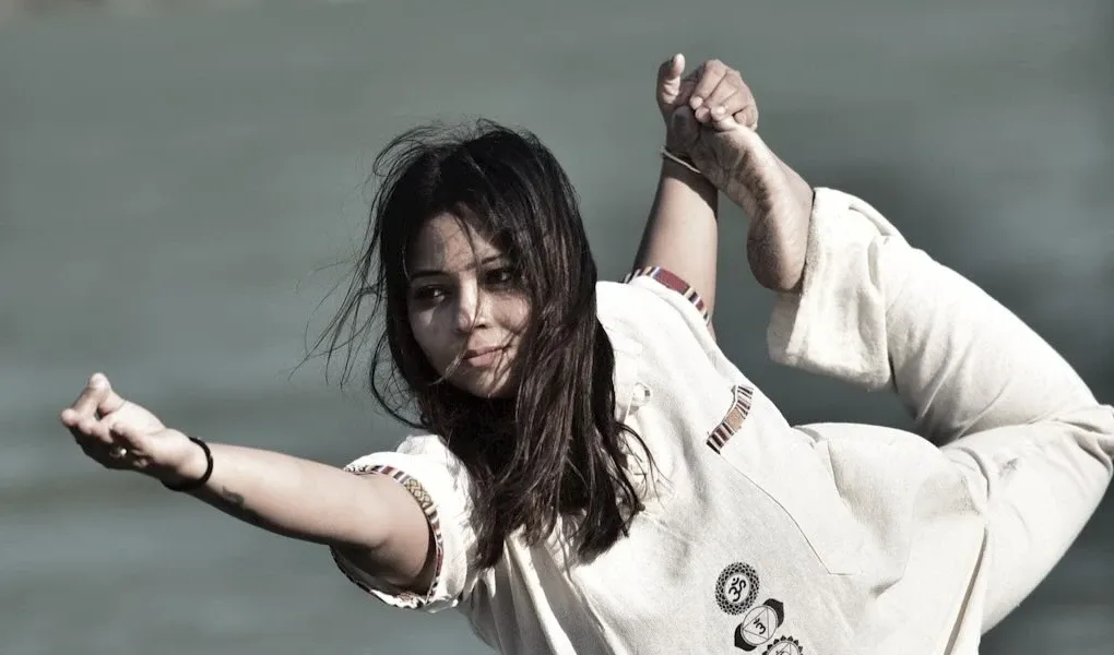 Anupriya holding a standing dancer pose by the water, her hair lifted by the wind