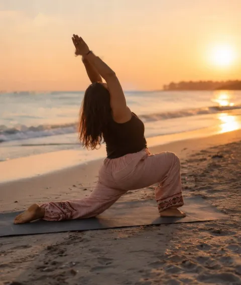 Woman in a low lunge with arms raised, practising yoga on a beach at sunset