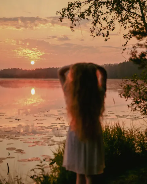A woman with long hair seen from behind, standing by a calm lake at sunset