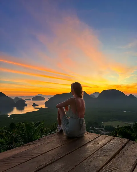 A person seated on a wooden deck, seen from behind, looking at a vivid orange sunrise over distant hills