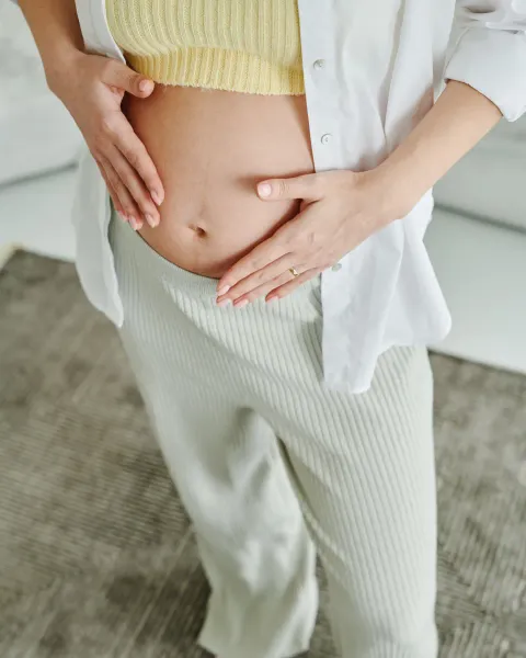 A pregnant woman in a white shirt and pale trousers resting her hands on her belly