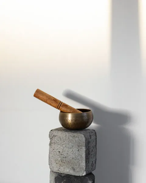 A small bronze singing bowl with a wooden mallet resting in it, on a stone block against a pale wall