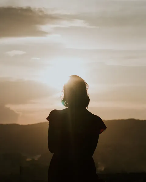 The silhouette of a person with their back to the camera, facing a hazy sunrise over low hills